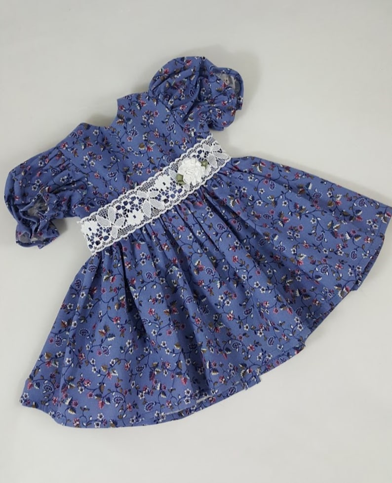 May include: A blue and white floral print dress with a white lace trim and a white flower accent. The dress has puffed sleeves and a gathered skirt.