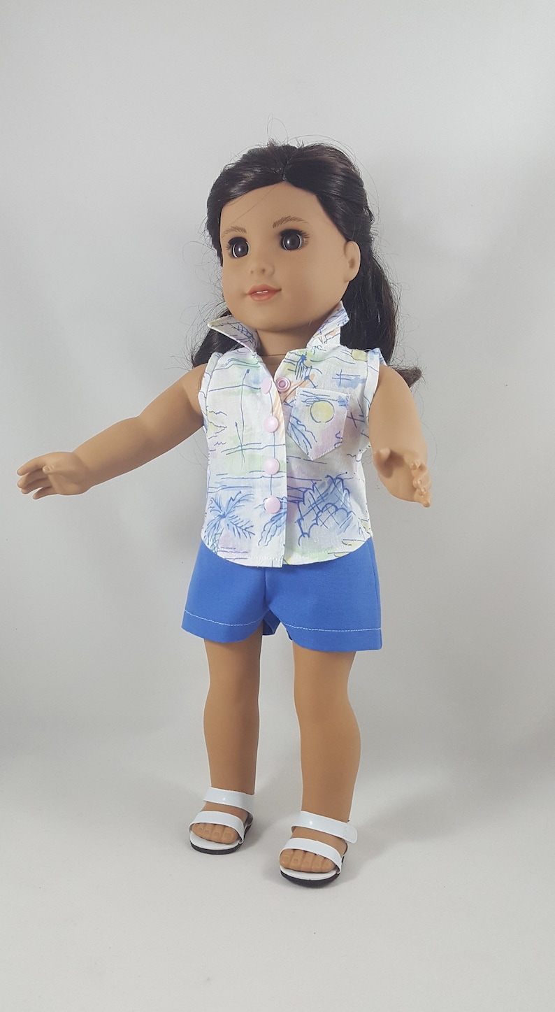 Peut inclure: Une poup&eacute;e portant une chemise blanche sans manches avec un imprim&eacute; palmier et un short bleu. La poup&eacute;e a les cheveux bruns et les yeux bruns et porte des sandales blanches.