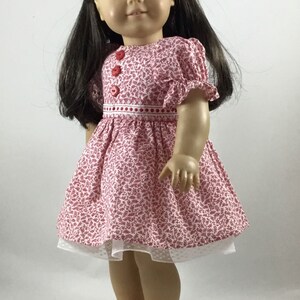 May include: A doll wearing a pink and white floral dress with red buttons and a white lace trim. The doll has brown hair and green eyes and is wearing red shoes.