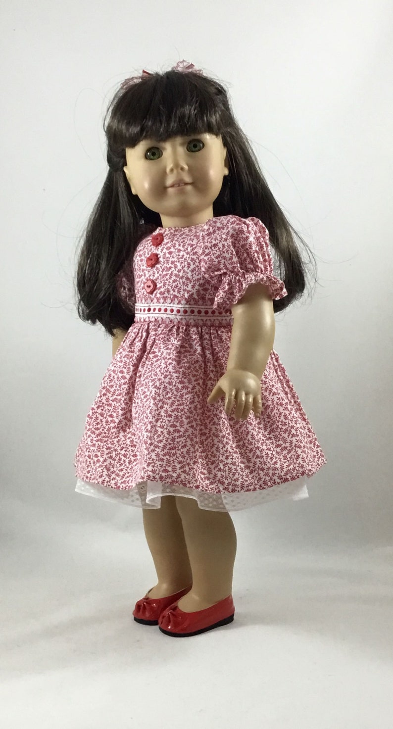 May include: A doll wearing a pink and white floral dress with red buttons and a white lace trim. The doll has brown hair and green eyes and is wearing red shoes.