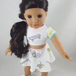 May include: A doll wearing a white crop top and skirt set with a skateboard and bicycle print. The top has a light blue and yellow sleeve. The doll has dark hair and is wearing purple sneakers.