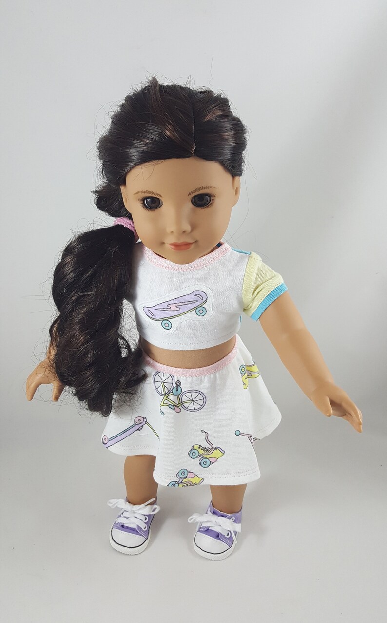 May include: A doll wearing a white crop top and skirt set with a skateboard and bicycle print. The top has a light blue and yellow sleeve. The doll has dark hair and is wearing purple sneakers.