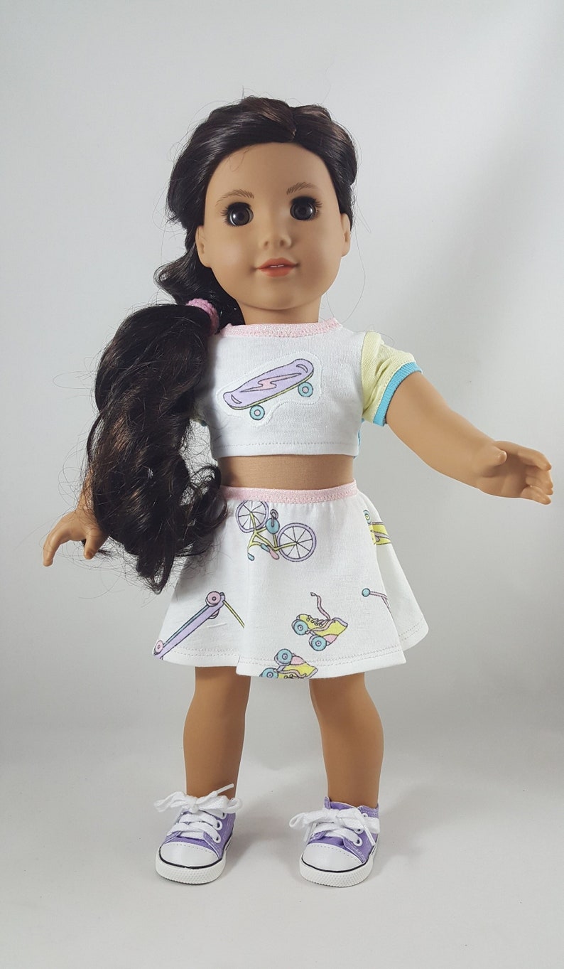 May include: A doll wearing a white crop top and skirt set with pastel skateboard and roller skate graphics. The top has yellow and blue accents. The doll has brown hair and is wearing purple sneakers.