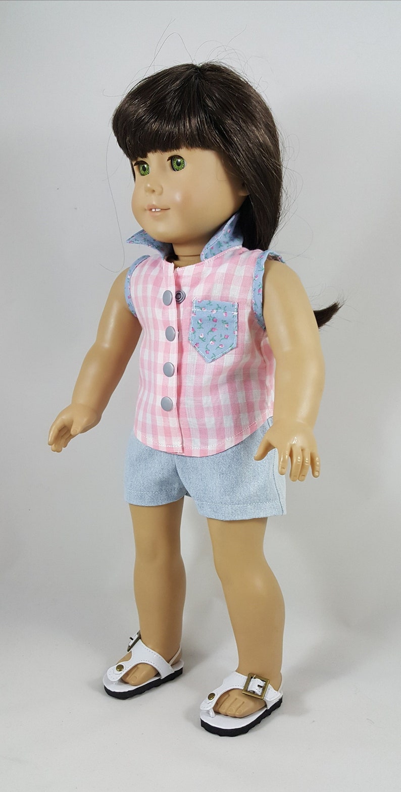 Peut inclure: Une poup&eacute;e portant une chemise sans manches en vichy rose et blanc avec une poche florale et un short en jean bleu. La poup&eacute;e a les cheveux bruns et les yeux verts et porte des sandales blanches avec des boucles dor&eacute;es.