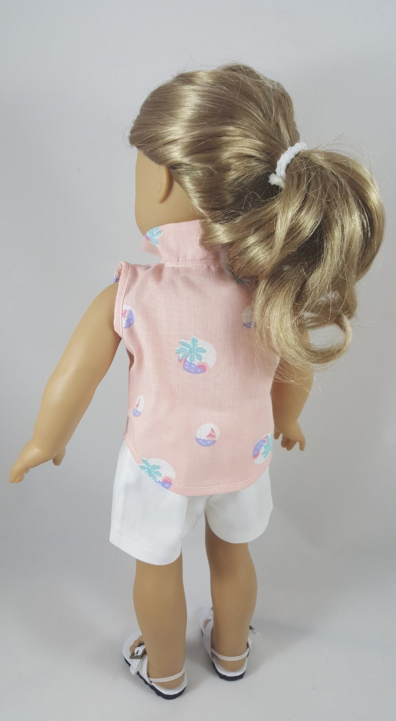 Peut inclure: Une poup&eacute;e portant une chemise rose sans manches avec un imprim&eacute; palmier et planche de surf et un short blanc. La poup&eacute;e a les cheveux blonds tir&eacute;s en arri&egrave;re en queue de cheval.