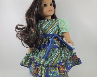 May include: A doll with long brown hair wearing a dress featuring a blue, green, and yellow paisley pattern with light green puff sleeves and a blue waist tie. The doll is wearing gold-colored sandals and has a light skin tone and brown eyes.
