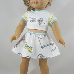 May include: A doll wearing a white crop top with roller skate patches and a matching skirt with skateboards, scooters, and bicycles. The top has yellow sleeves and blue trim. The doll is wearing white sneakers.