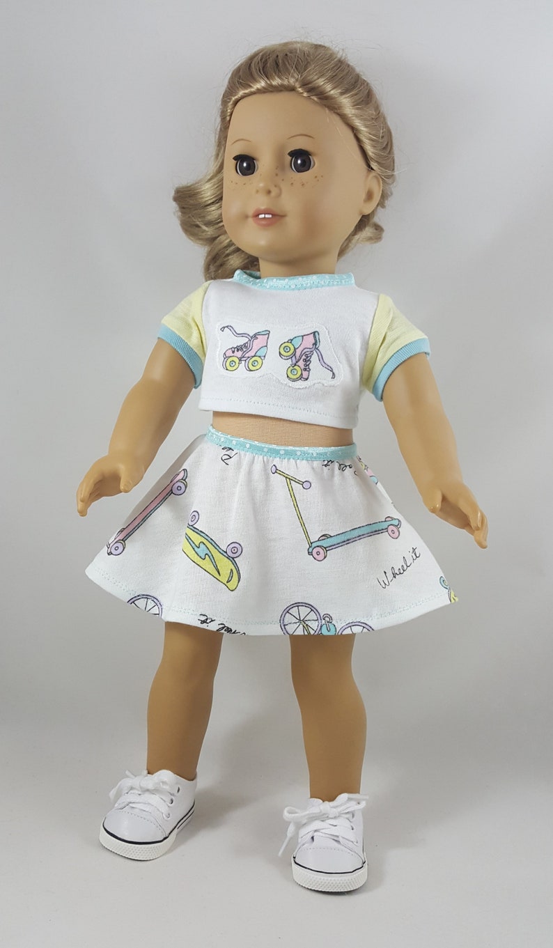 May include: A doll wearing a white crop top with roller skate patches and a matching skirt with skateboards, scooters, and bicycles. The top has yellow sleeves and blue trim. The doll is wearing white sneakers.