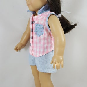 Peut inclure: Une poup&eacute;e portant une chemise sans manches en vichy rose et blanc avec une poche florale bleue et un short en jean bleu. La poup&eacute;e porte des sandales blanches avec une boucle marron.