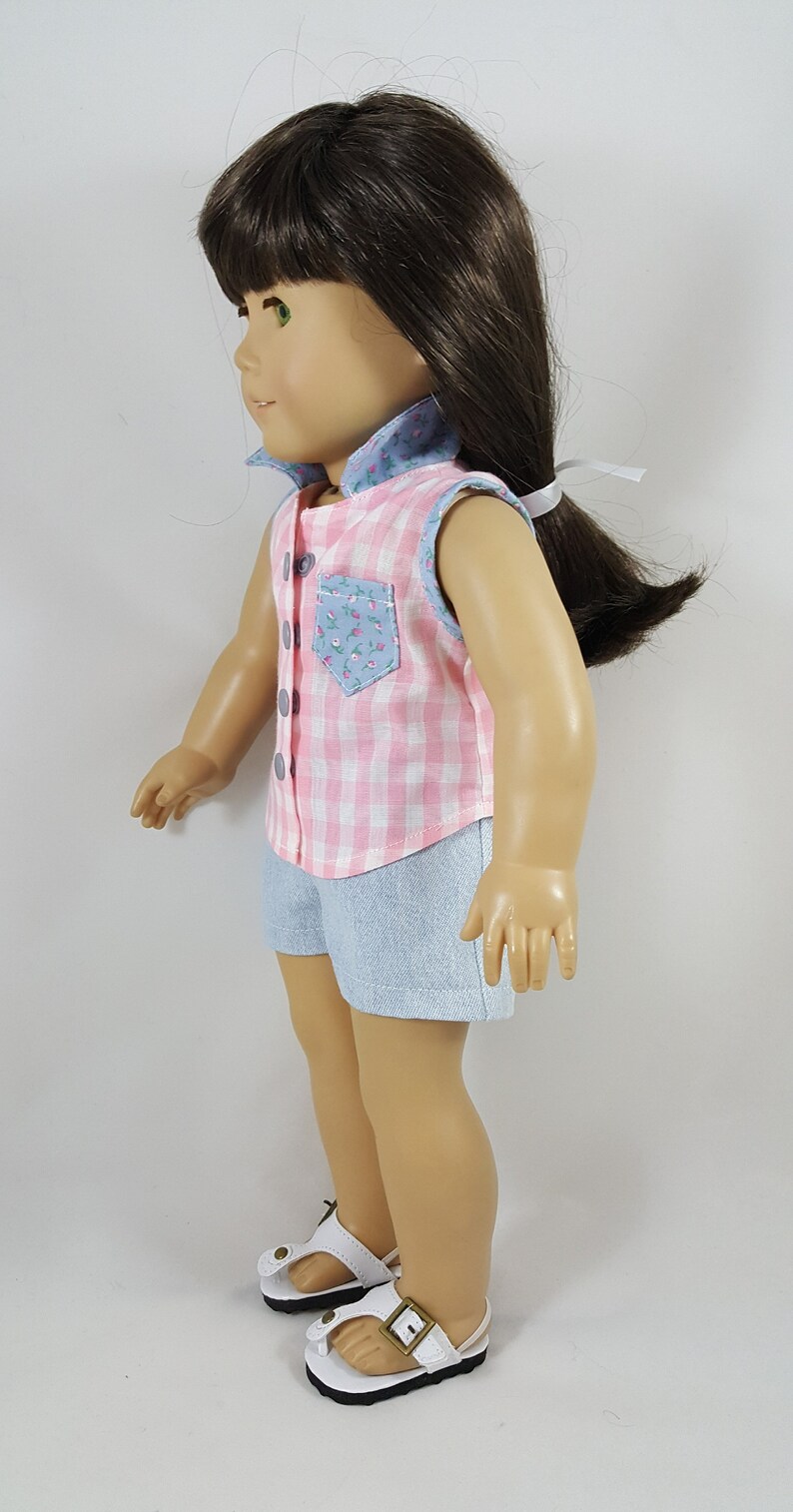 Peut inclure: Une poup&eacute;e portant une chemise sans manches en vichy rose et blanc avec une poche florale bleue et un short en jean bleu. La poup&eacute;e porte des sandales blanches avec une boucle marron.