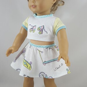 May include: A doll wearing a white crop top and skirt set with a roller skate and scooter pattern. The top has yellow and blue trim, and the skirt has a matching trim. The doll is also wearing white sneakers.