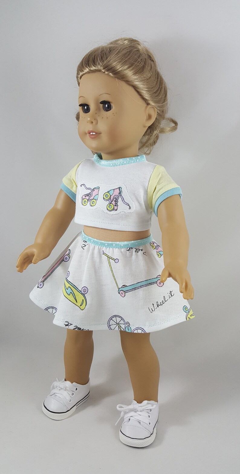 May include: A doll wearing a white crop top and skirt set with a roller skate and scooter pattern. The top has yellow and blue trim, and the skirt has a matching trim. The doll is also wearing white sneakers.
