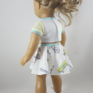 May include: A doll wearing a white crop top with yellow sleeves and blue trim, paired with a matching skirt. The skirt features colorful illustrations of skateboards and bicycles. The doll is also wearing white sneakers.