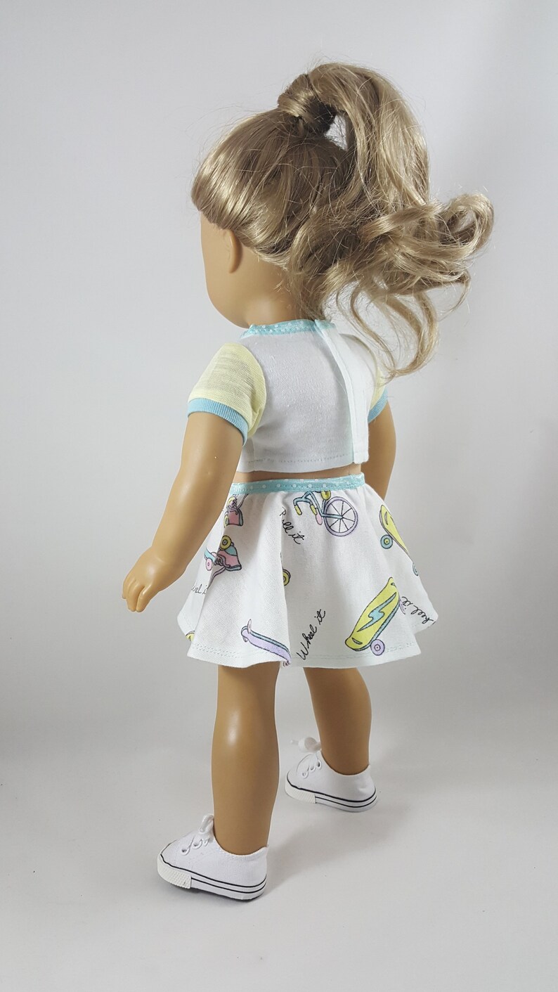 May include: A doll wearing a white crop top with yellow sleeves and blue trim, paired with a matching skirt. The skirt features colorful illustrations of skateboards and bicycles. The doll is also wearing white sneakers.