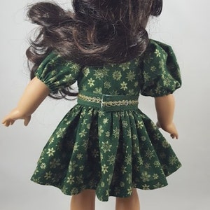 Op de afbeelding: Een groene jurk met gouden sneeuwvlokkenprint en een gouden riem. De jurk heeft pofmouwen en een volle rok. De pop draagt zwarte schoenen.