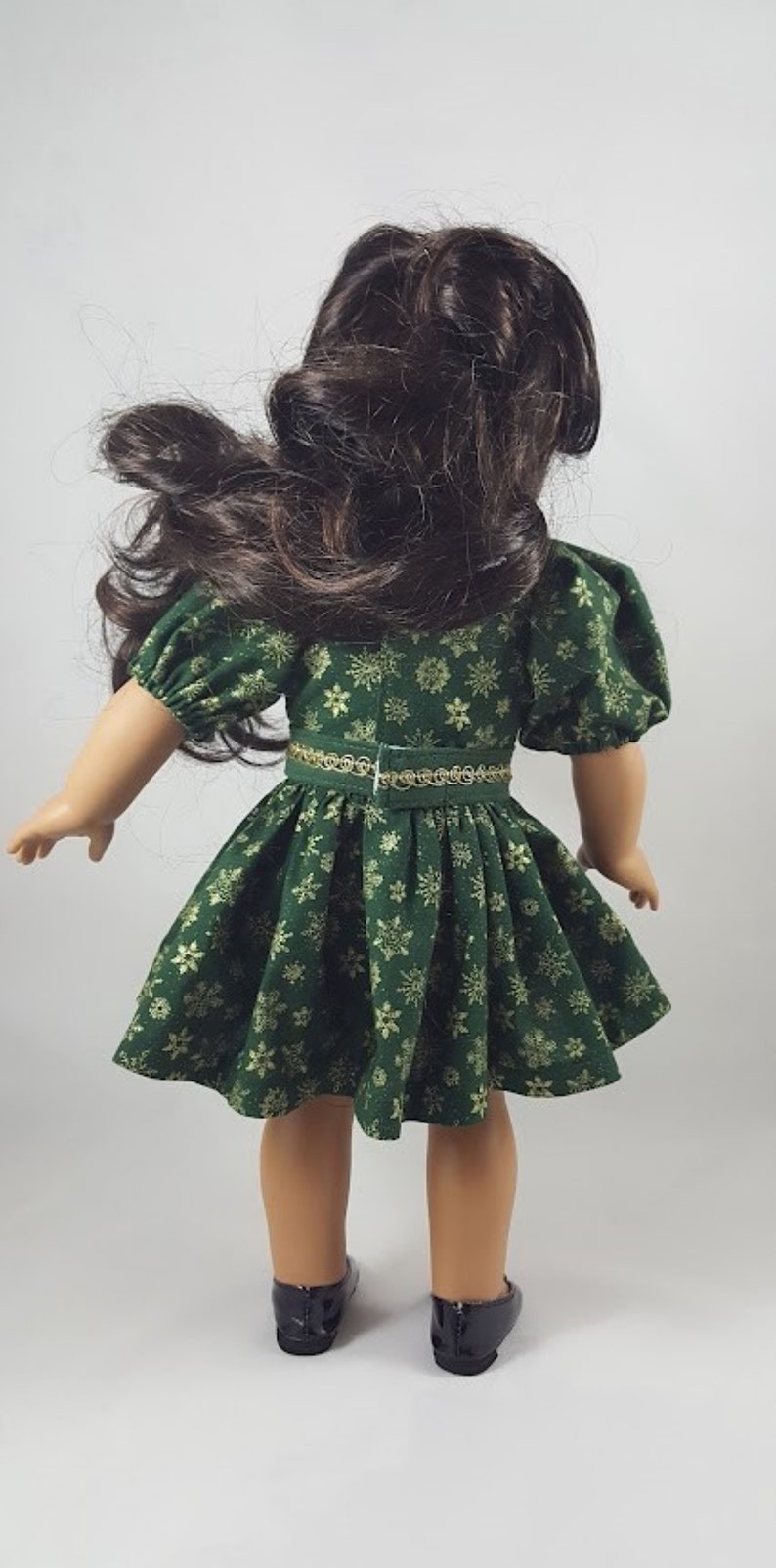 Op de afbeelding: Een groene jurk met gouden sneeuwvlokkenprint en een gouden riem. De jurk heeft pofmouwen en een volle rok. De pop draagt zwarte schoenen.