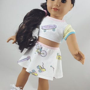 May include: A doll wearing a white crop top and skirt set with pastel skateboard and bicycle graphics. The top has a yellow and blue sleeve. The skirt and top have pink trim. The doll is wearing purple sneakers with white laces.