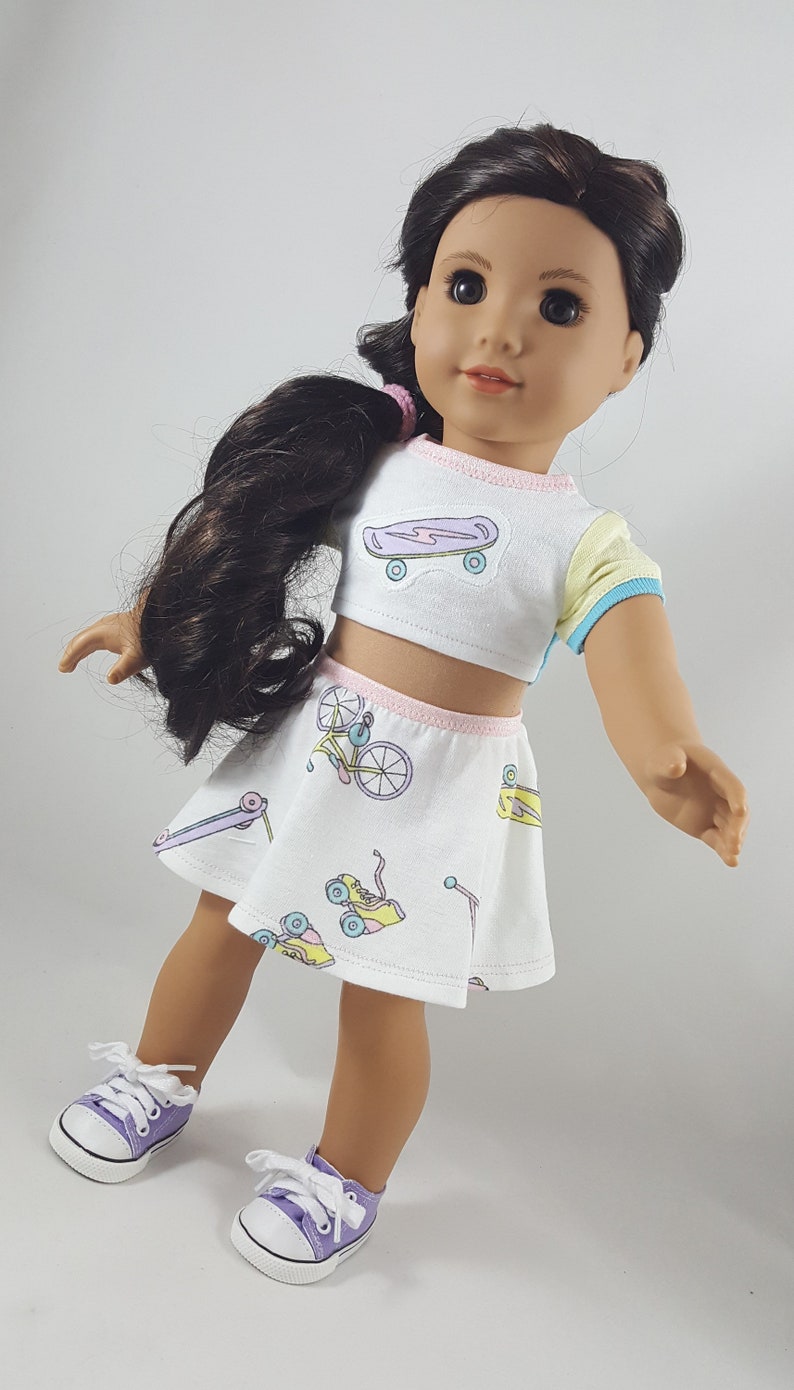 May include: A doll wearing a white crop top and skirt set with pastel skateboard and bicycle graphics. The top has a yellow and blue sleeve. The skirt and top have pink trim. The doll is wearing purple sneakers with white laces.