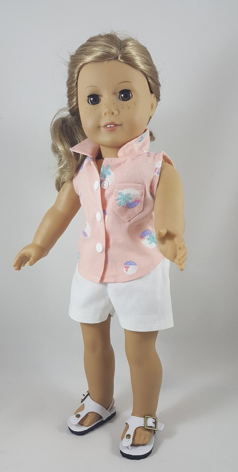 Peut inclure: Une poup&eacute;e portant une chemise rose sans manches avec un imprim&eacute; palmier et un short blanc. La poup&eacute;e a les cheveux blonds et les yeux bruns. La poup&eacute;e porte des sandales blanches avec une boucle.
