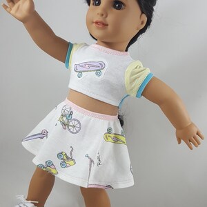 May include: A doll wearing a white crop top and skirt set with a skateboard and roller skate print. The top has yellow and blue sleeves. The skirt has a pink waistband. The doll is wearing purple sneakers.
