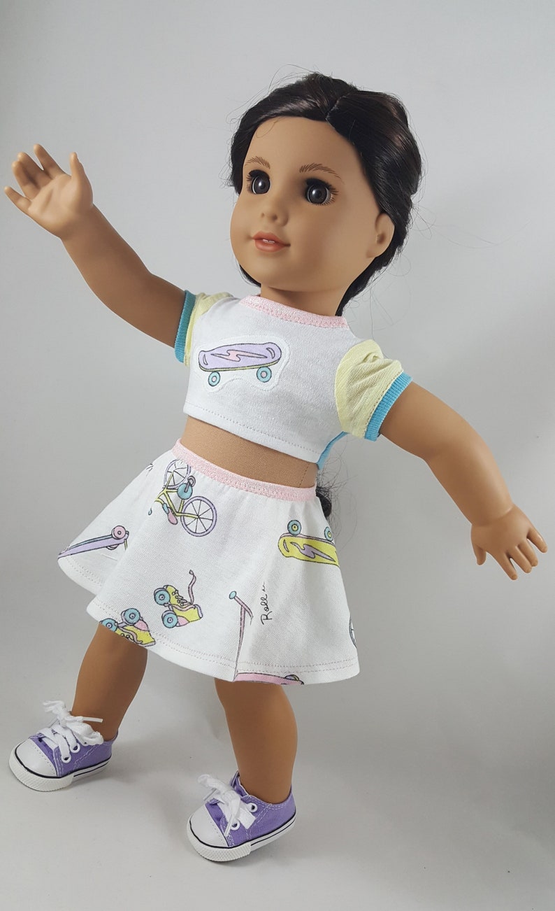 May include: A doll wearing a white crop top and skirt set with a skateboard and roller skate print. The top has yellow and blue sleeves. The skirt has a pink waistband. The doll is wearing purple sneakers.