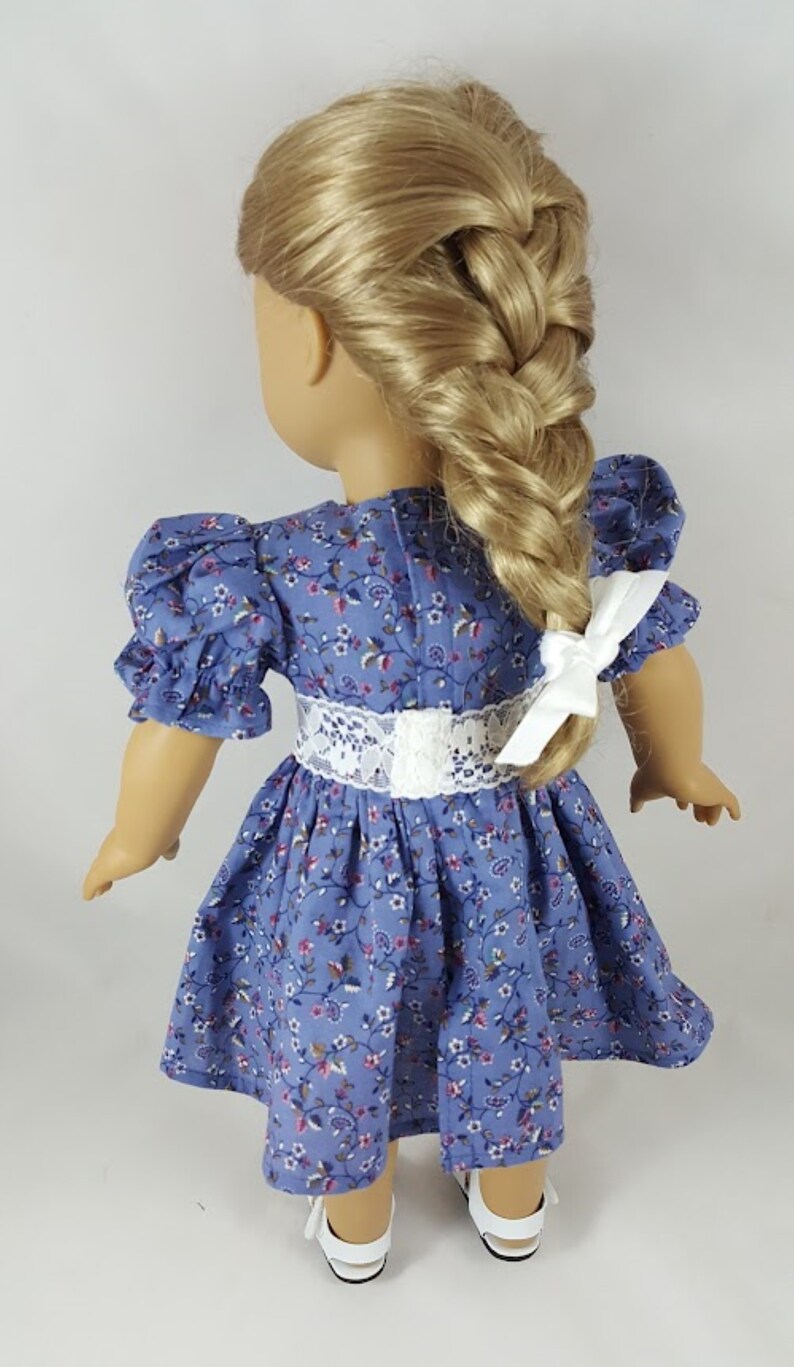 May include: A blue floral dress with a white lace waistband and puffed sleeves. The dress is worn by a doll with blonde hair styled in a braid.