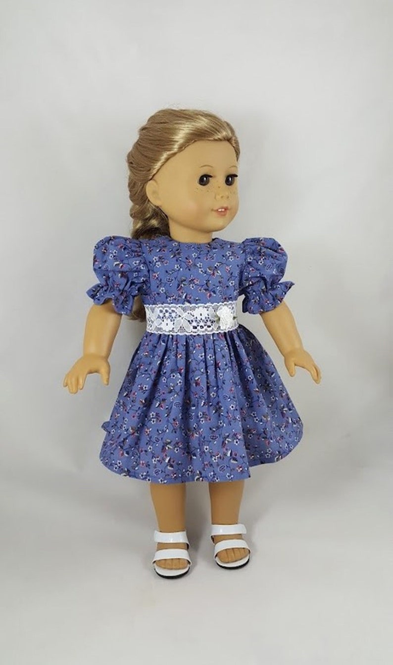 May include: A doll wearing a blue and white floral dress with a white lace belt and white sandals.