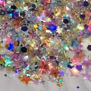 May include: A close-up of a pile of iridescent glitter in various shapes, including stars and hexagons. The glitter is a mix of colors, including pink, blue, yellow, and orange, creating a sparkling effect. Ideal for crafts and decorations.