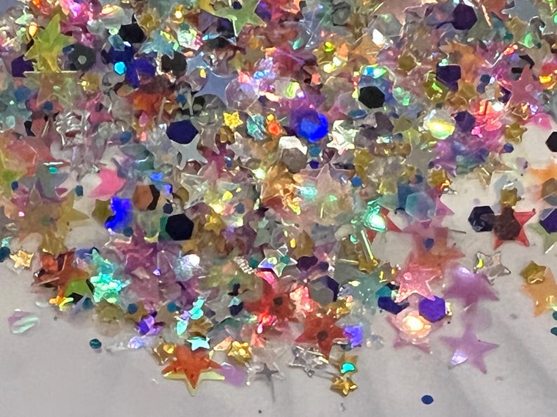 May include: A close-up of a pile of iridescent glitter in various shapes, including stars and hexagons. The glitter is a mix of colors, including pink, blue, yellow, and orange, creating a sparkling effect. Ideal for crafts and decorations.