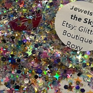 May include: A close-up of a bag filled with colorful glitter. The glitter includes star and hexagonal shapes in various colors, including pink, blue, green, and gold. The bag has a white label with the text "Jewels in the Sky Etsy: Glitter Boutique Roxy."