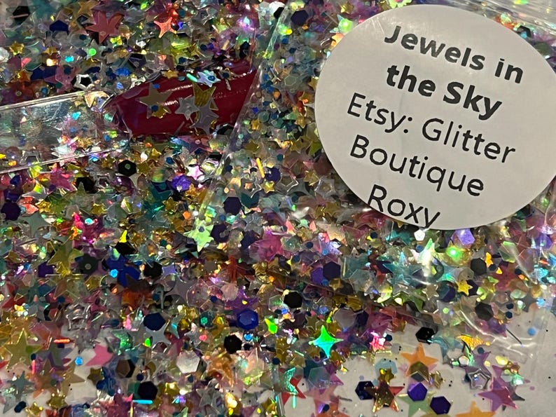 May include: A close-up of a bag filled with colorful glitter. The glitter includes star and hexagonal shapes in various colors, including pink, blue, green, and gold. The bag has a white label with the text "Jewels in the Sky Etsy: Glitter Boutique Roxy."