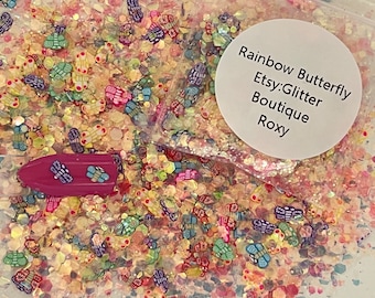 May include: A close-up of a clear plastic bag filled with colorful glitter shaped like butterflies. The glitter is a mix of gold, pink, blue, green, and purple. A white label on the bag reads "Rainbow Butterfly Etsy:Glitter Boutique Roxy".