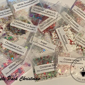 May include: A collection of small, clear plastic bags filled with various colors and shapes of glitter. Each bag is labeled with a Christmas-themed name, such as "Christmas Lights LG" and "Winter Pink." The words "Sample Pack Christmas" are at the bottom.