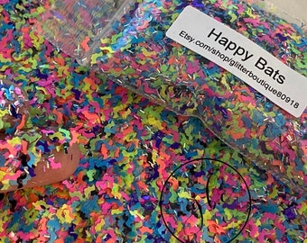 May include: A close-up of colorful glitter in the shape of bats. The glitter is a mix of neon pink, yellow, blue, and purple. A small bag of glitter is visible with the text "Happy Bats" and a shop URL.