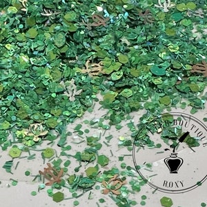 May include: Green, gold, and iridescent glitter mix with various shapes, including small circles, hexagons, and leaves. The glitter is scattered on a white surface. A round sticker with the text "Glitter Boutique Roxy" is in the lower right corner.
