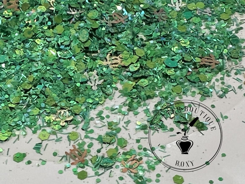 May include: Green, gold, and iridescent glitter mix with various shapes, including small circles, hexagons, and leaves. The glitter is scattered on a white surface. A round sticker with the text "Glitter Boutique Roxy" is in the lower right corner.