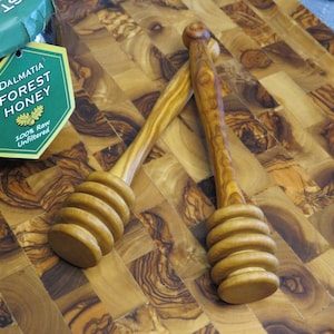 May include: Two wooden honey dippers with a rounded handle and a spiral design. The dippers are on a wooden cutting board with a natural wood grain pattern.