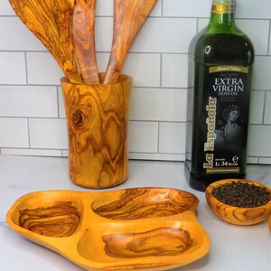 May include: A set of olive wood kitchen utensils, including a serving spoon, a slotted spoon, and a spatula, are displayed in a wooden holder. A bottle of extra virgin olive oil and a small olive wood bowl filled with black peppercorns are also shown.