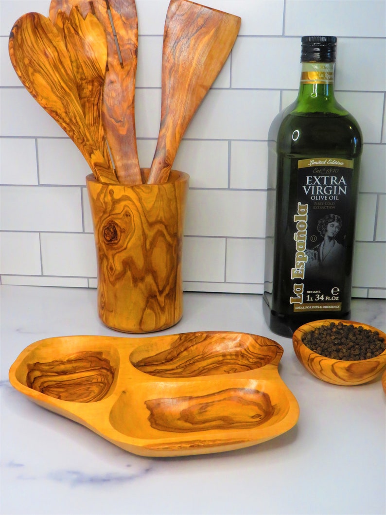 May include: A set of olive wood kitchen utensils, including a serving spoon, a slotted spoon, and a spatula, are displayed in a wooden holder. A bottle of extra virgin olive oil and a small olive wood bowl filled with black peppercorns are also shown.