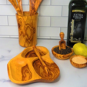 May include: Olive wood serving dish with three compartments and a small fork. The dish is filled with black peppercorns, salt, and a lemon wedge. There is also a bottle of olive oil and a wooden utensil holder with three wooden spoons.
