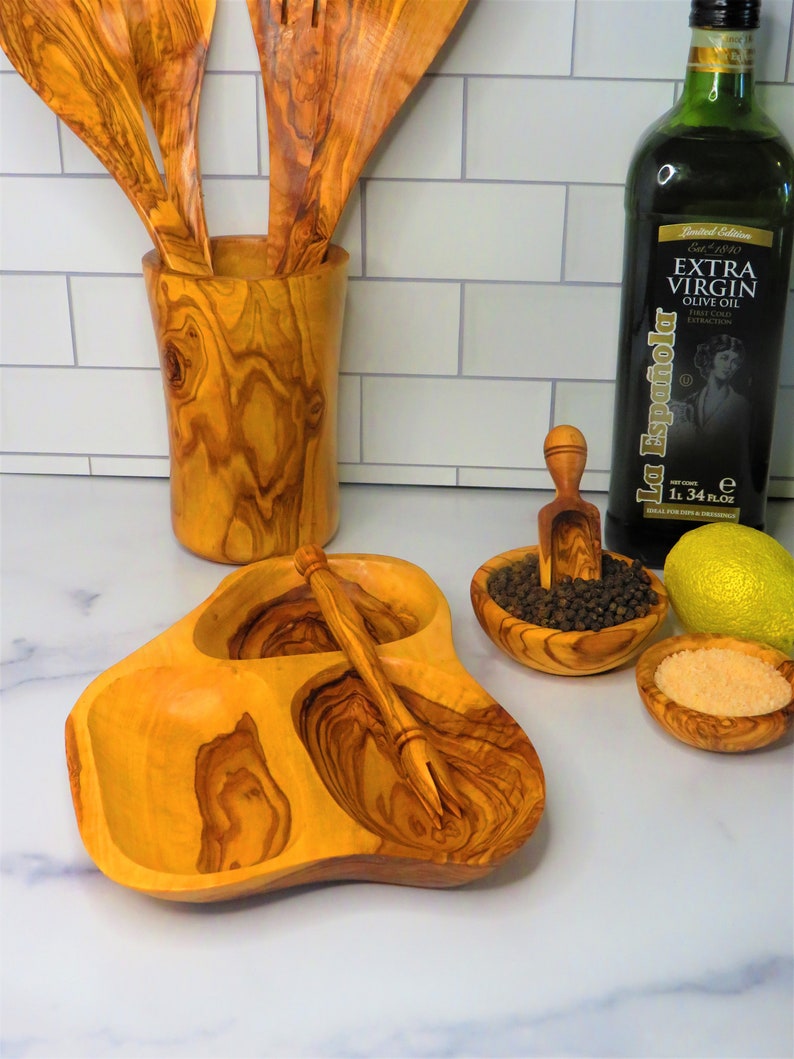 May include: Olive wood serving dish with three compartments and a small fork. The dish is filled with black peppercorns, salt, and a lemon wedge. There is also a bottle of olive oil and a wooden utensil holder with three wooden spoons.