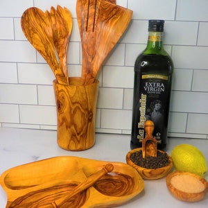 May include: Olive wood serving tray with three compartments, a small olive wood spoon, and a small olive wood scoop.  The tray is filled with black peppercorns, salt, and a lemon.  A bottle of olive oil and a wooden utensil holder with olive wood spoons are in the background.