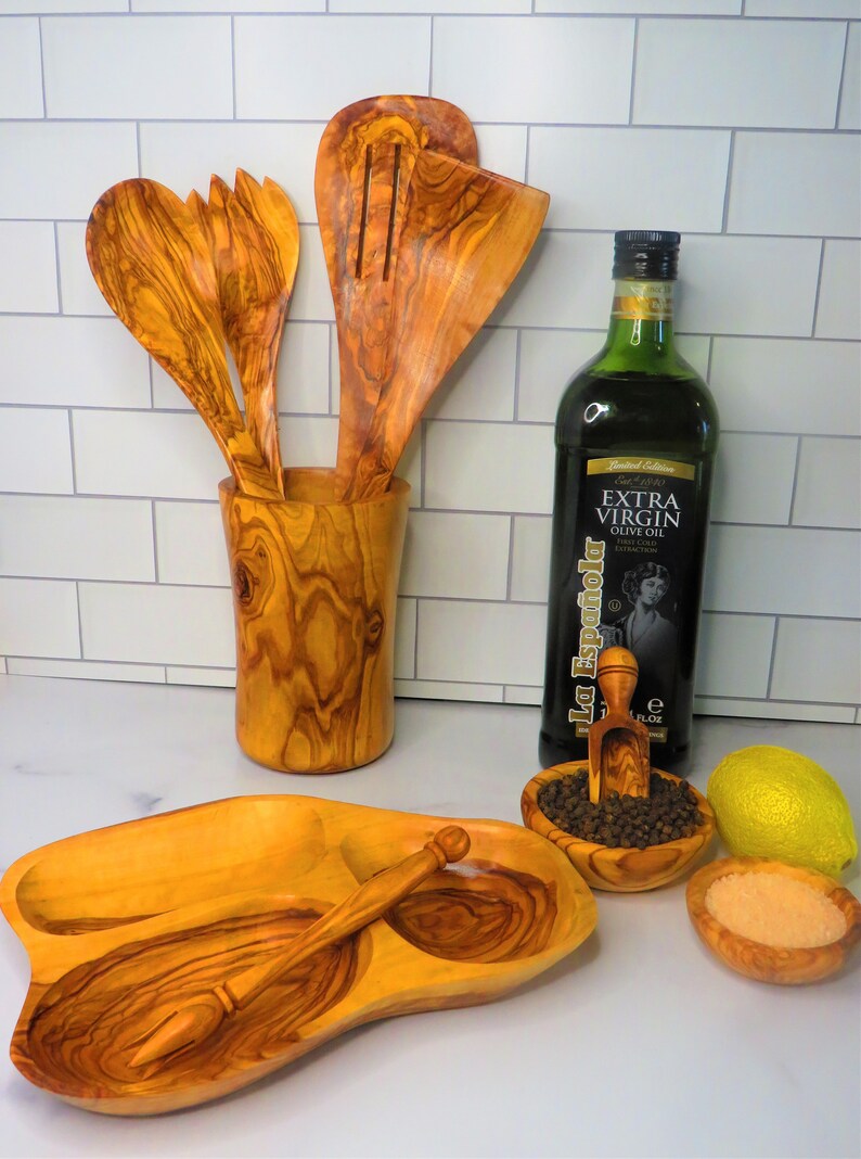 May include: Olive wood serving tray with three compartments, a small olive wood spoon, and a small olive wood scoop.  The tray is filled with black peppercorns, salt, and a lemon.  A bottle of olive oil and a wooden utensil holder with olive wood spoons are in the background.