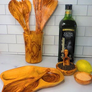 May include: A set of olive wood kitchen utensils, including a serving spoon, a slotted spoon, a fork, and a small spoon, are displayed in a wooden holder. A bottle of olive oil, a small bowl of black peppercorns, a small bowl of salt, and a lemon are also shown on a white tiled countertop.