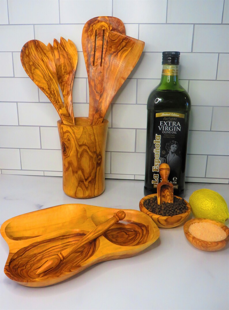 May include: A set of olive wood kitchen utensils, including a serving spoon, a slotted spoon, a fork, and a small spoon, are displayed in a wooden holder. A bottle of olive oil, a small bowl of black peppercorns, a small bowl of salt, and a lemon are also shown on a white tiled countertop.