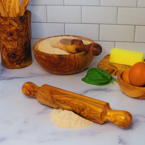 May include: A wooden rolling pin with a natural wood grain finish. The rolling pin is lying on a white surface with a small pile of flour next to it.