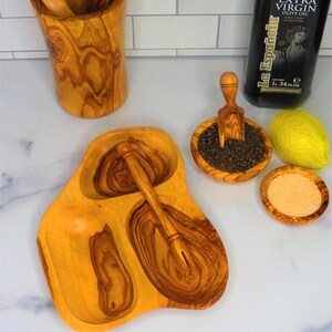 May include: Olive wood serving tray with three separate bowls, a small wooden spoon, and a scoop. The tray is on a white marble surface. There is a bottle of olive oil and a lemon in the background.