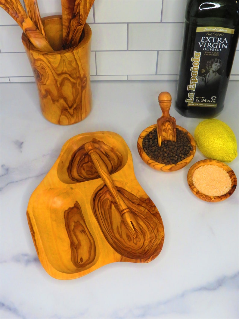 May include: Olive wood serving tray with three separate bowls, a small wooden spoon, and a scoop. The tray is on a white marble surface. There is a bottle of olive oil and a lemon in the background.