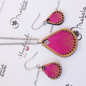 May include: A pink and brown wooden necklace and earrings set. The necklace has a teardrop-shaped pendant with a pink yarn tassel. The earrings are also teardrop-shaped with pink yarn tassels.