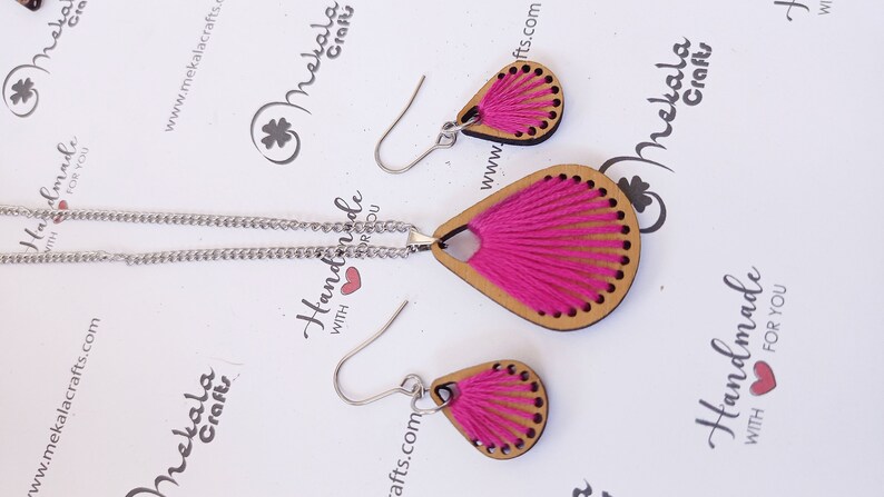 May include: A pink and brown wooden necklace and earrings set. The necklace has a teardrop-shaped pendant with a pink yarn tassel. The earrings are also teardrop-shaped with pink yarn tassels.